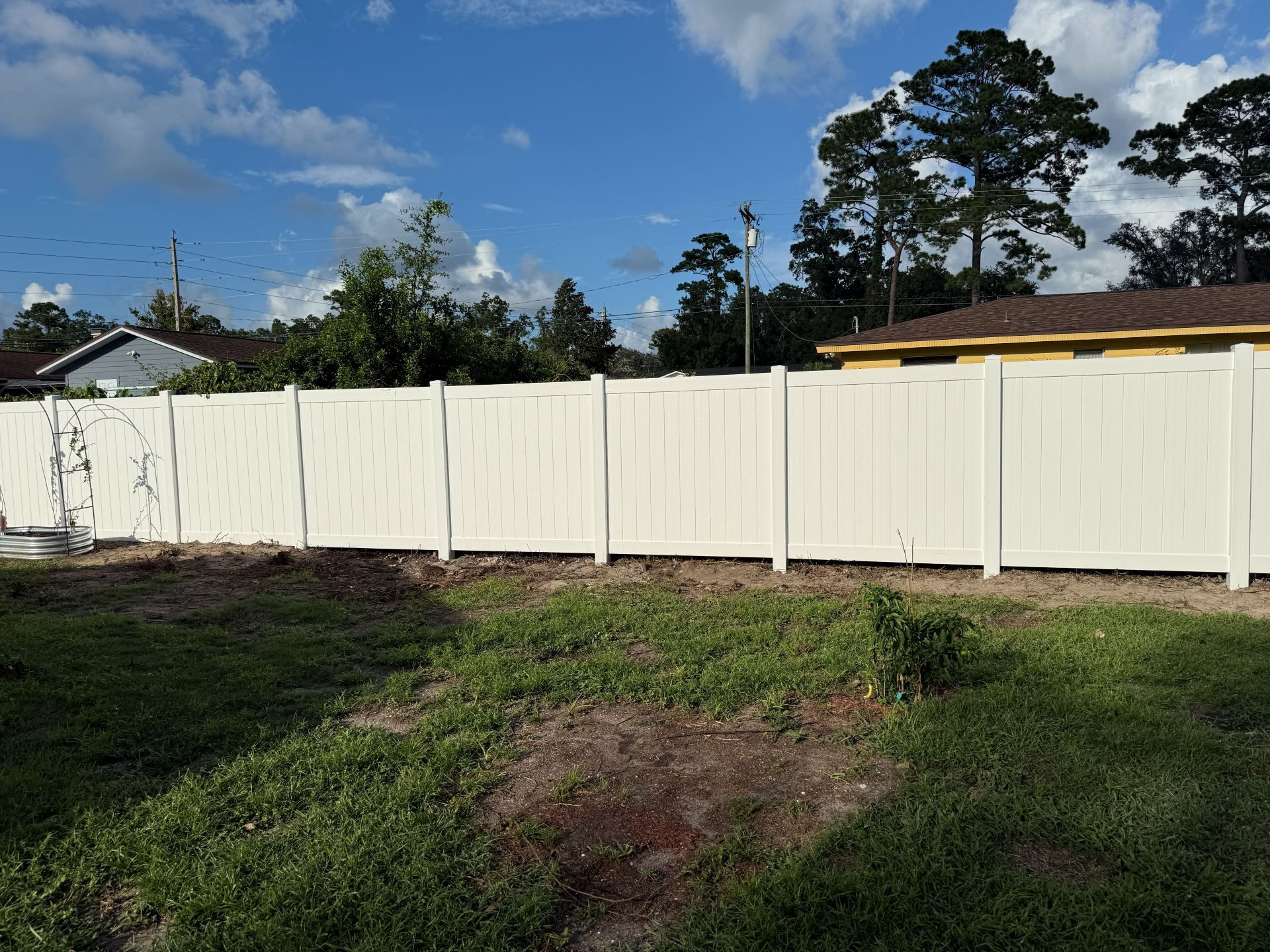 White fence along a grassy yard under a blue sky with clouds and trees behind it