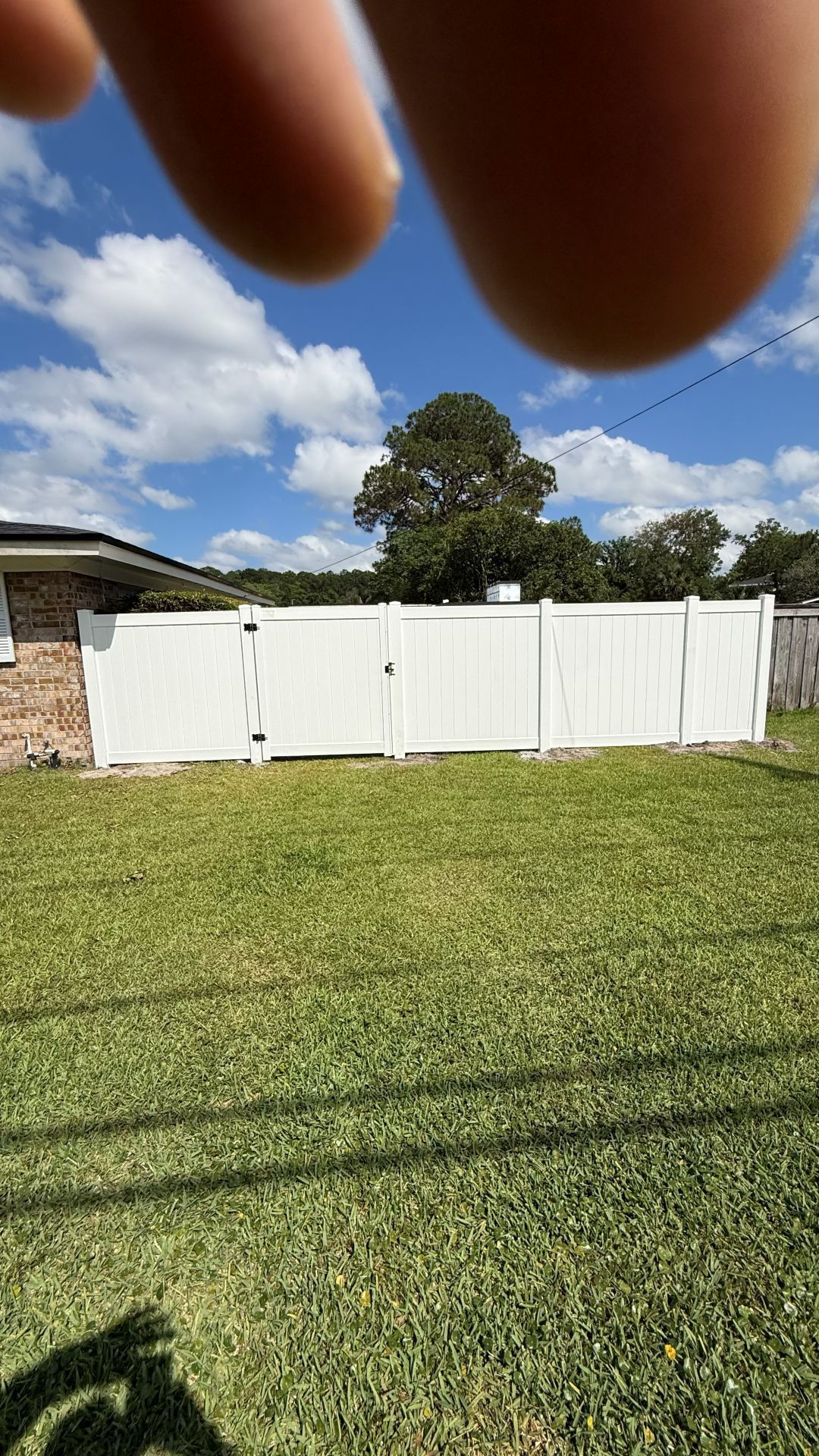 White fence in a grassy backyard under a partly cloudy sky, with fingers partly covering the top of the view.
