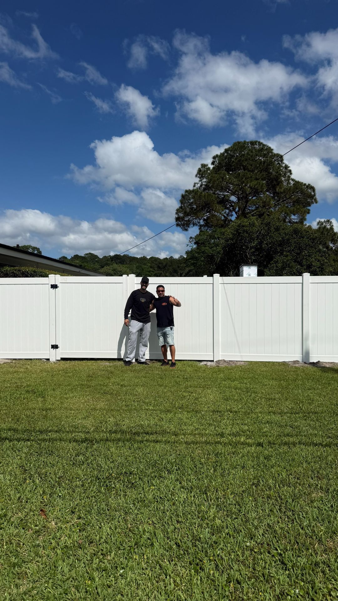 Two people standing by a white fence on a grassy field under a blue sky with clouds.