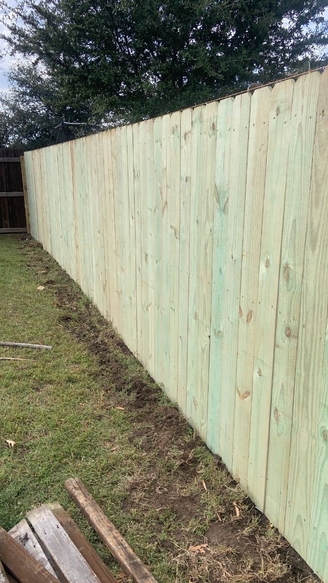 Freshly installed light-green wooden fence along a grassy yard, with a stack of lumber in the foreground.