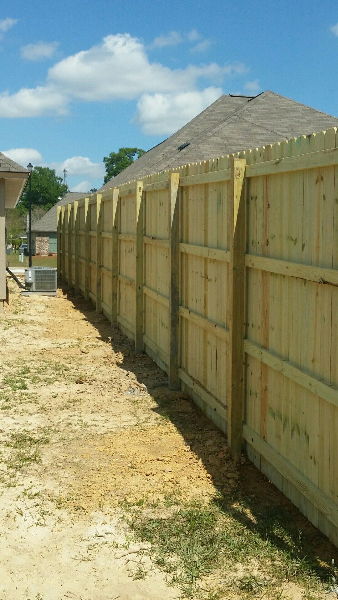 New wooden privacy fence along a dirt side yard under a blue sky with clouds