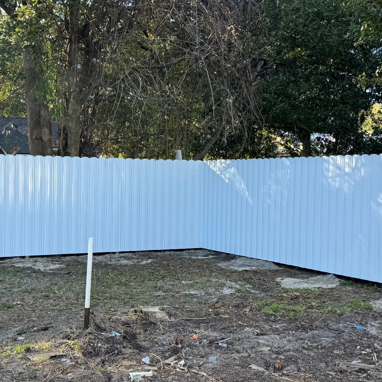 White corrugated metal fence around a bare dirt yard with trees in the background