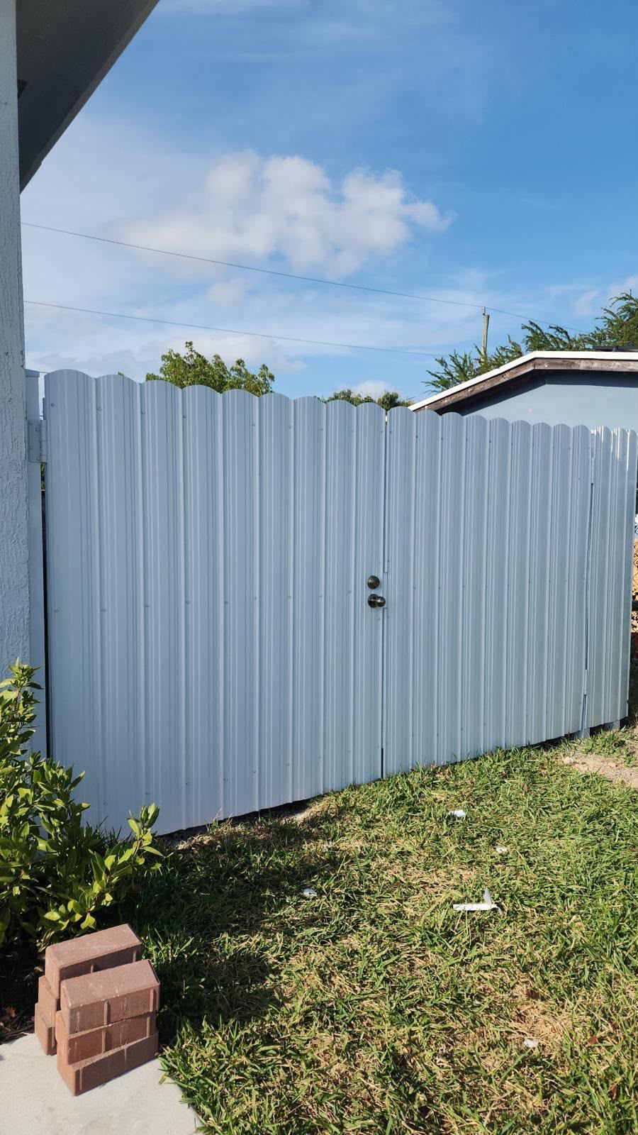 Corrugated metal fence gate beside grassy yard under blue sky, with a small stack of bricks in front