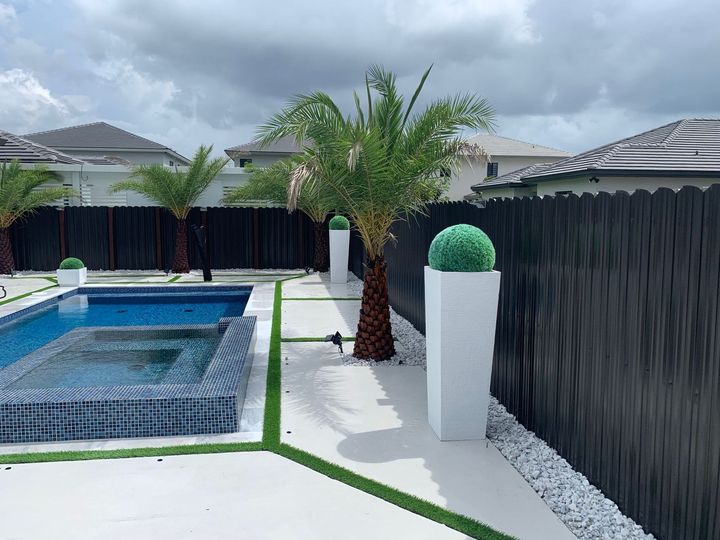 Backyard pool with palm trees, white planters, black fencing, and a narrow concrete walkway.