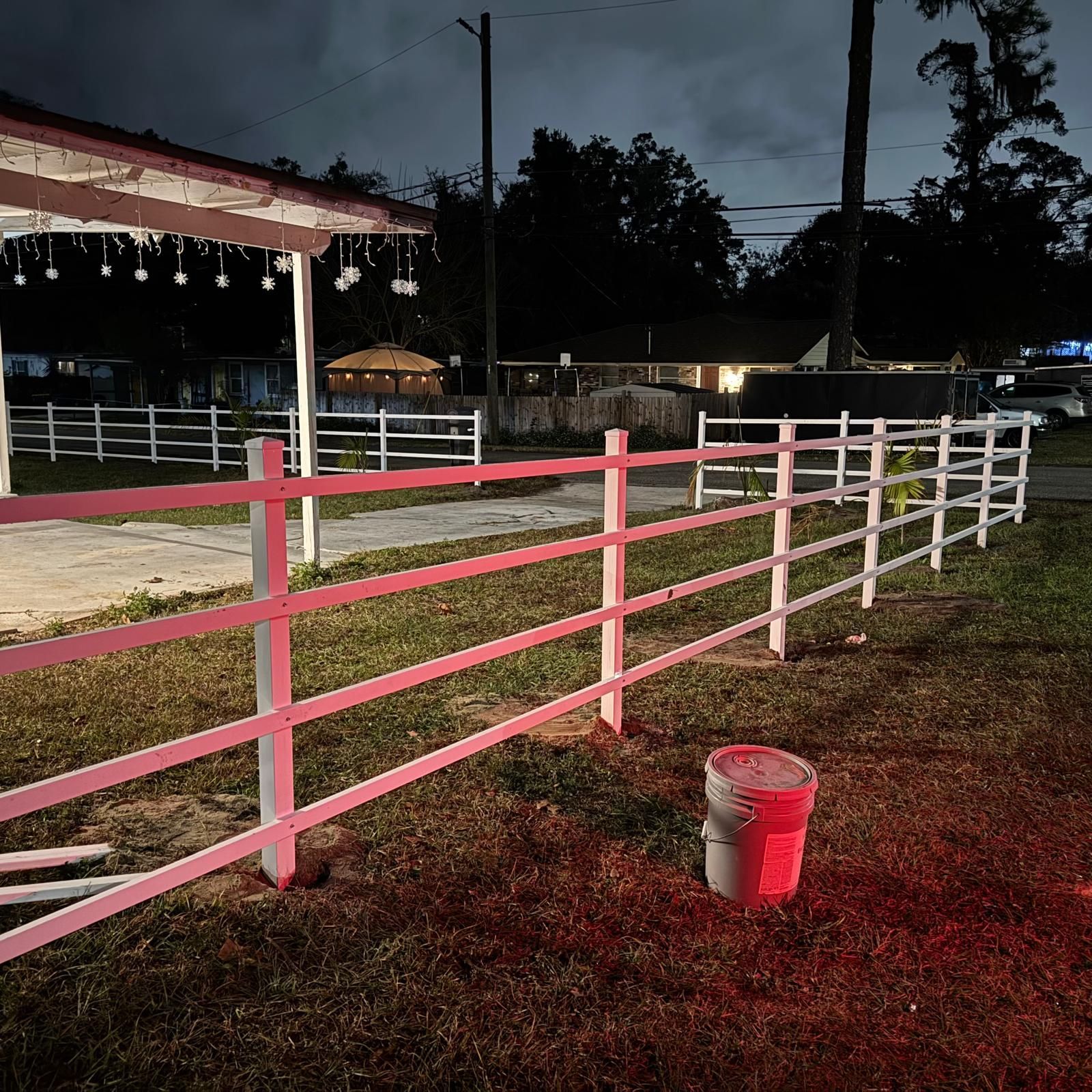 Pink and white fence by a barn at dusk, with a red bucket on the grass