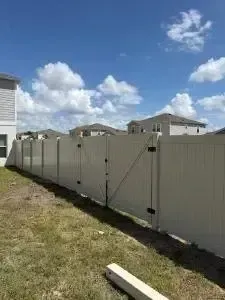 White vinyl fence lining a grassy yard under a blue sky with clouds, between suburban houses.