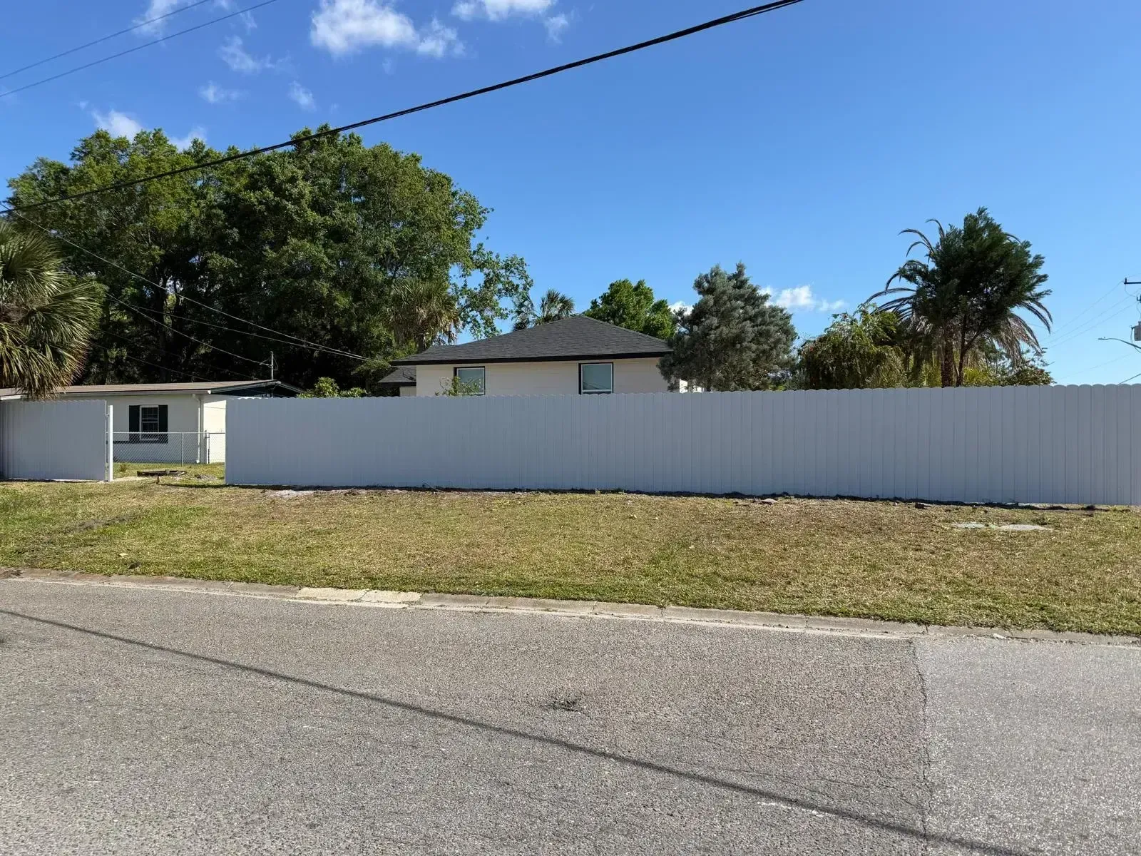 Suburban house behind a long white fence under a blue sky, with street and grass in front.