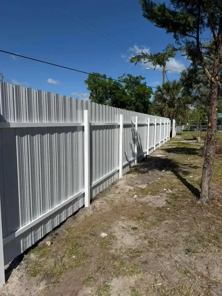 White vinyl fence along a dirt path under a blue sky, with trees and grass beside it.