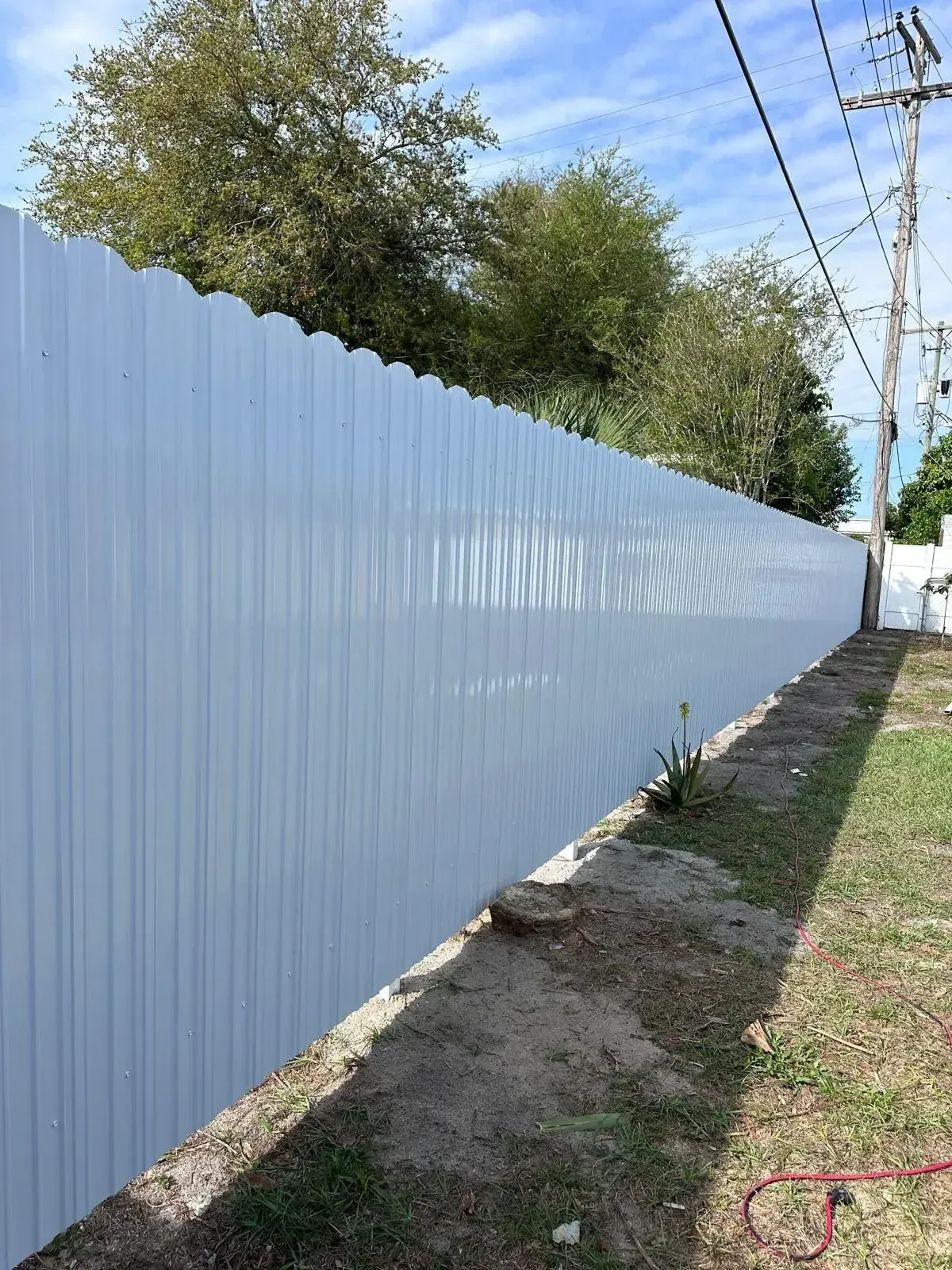 Long white corrugated fence beside a dirt path, with trees and utility lines along the side.