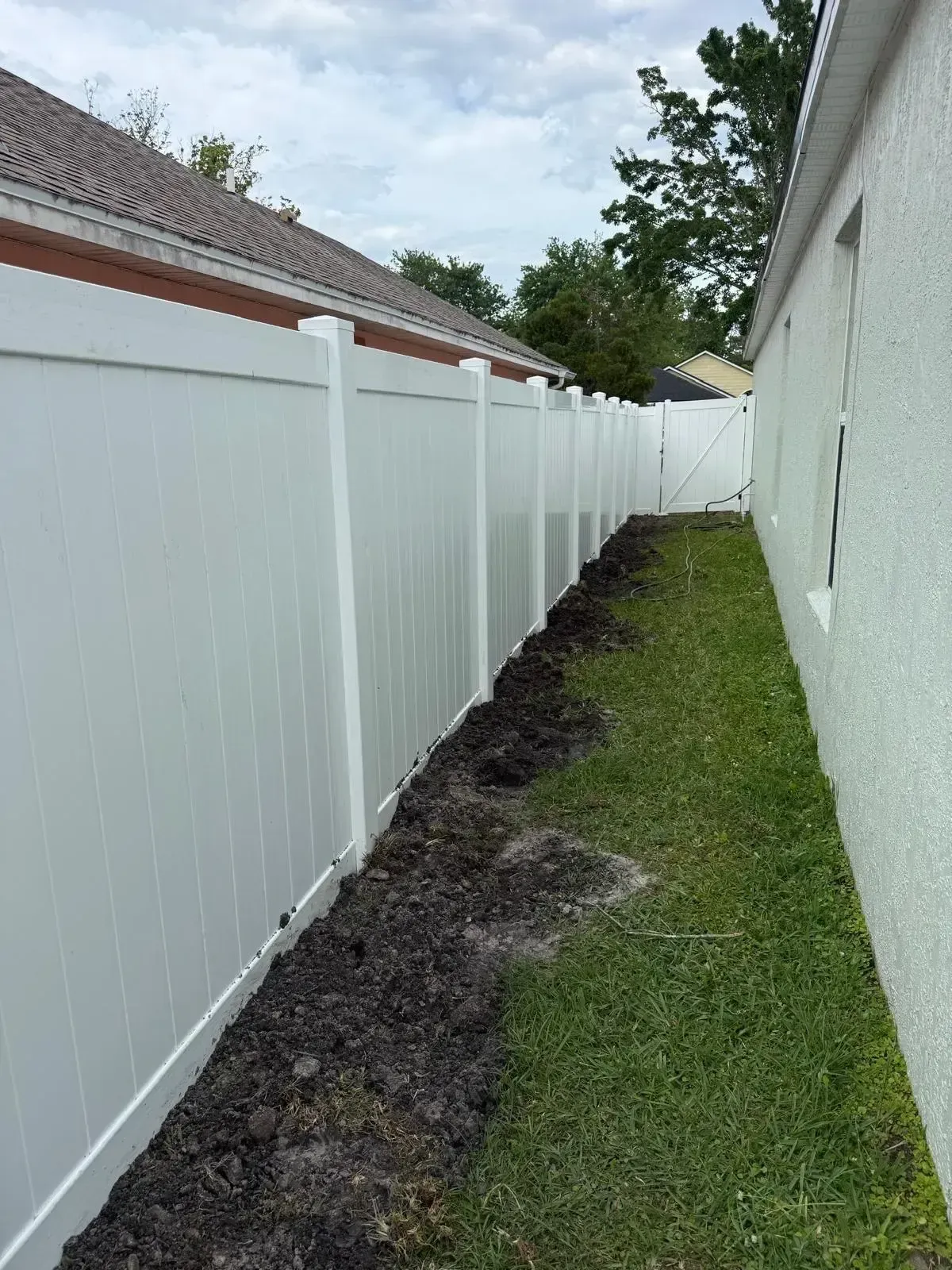 Narrow side yard with white vinyl fence, house wall, grass, and a dark mulched strip along the fence