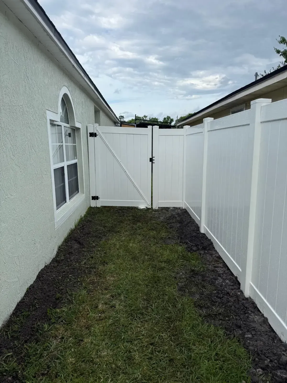 Narrow backyard side yard with white house wall, white fence, and a gate at the end.