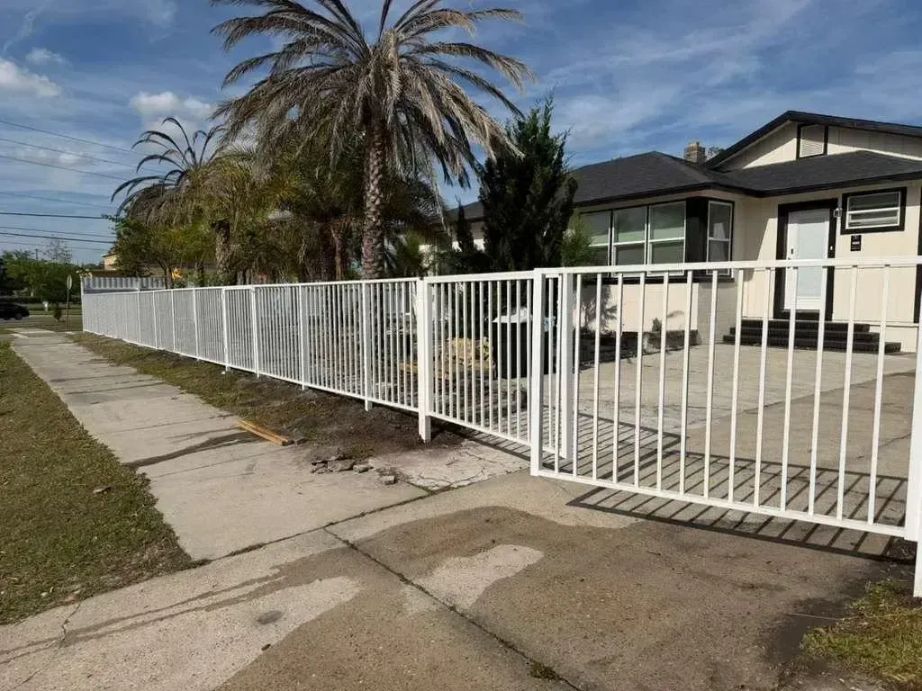 White metal fence along a residential sidewalk beside a house and tall palm tree, under a blue sky.