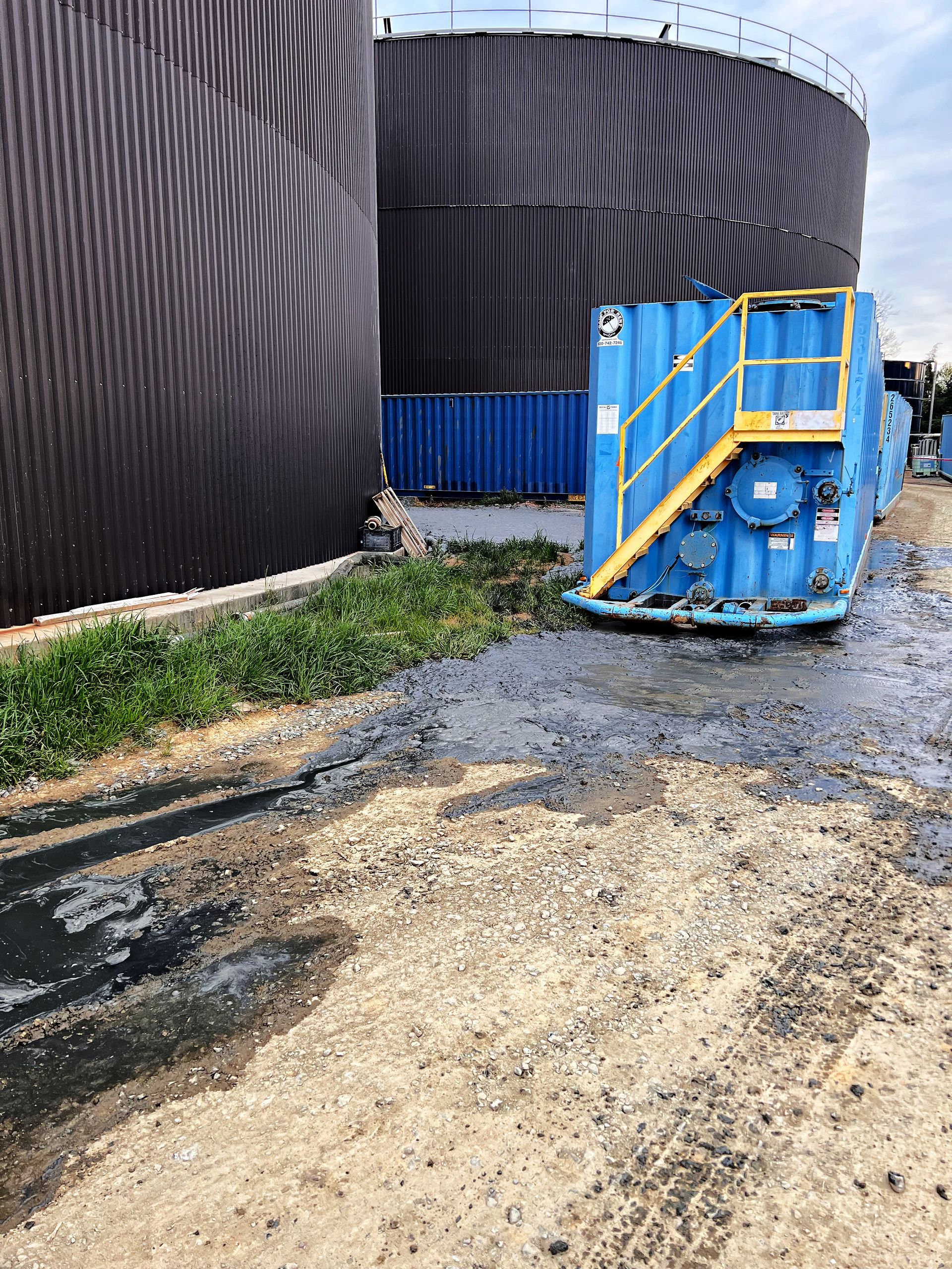 Blue industrial equipment beside large black storage tanks on a gravel lot