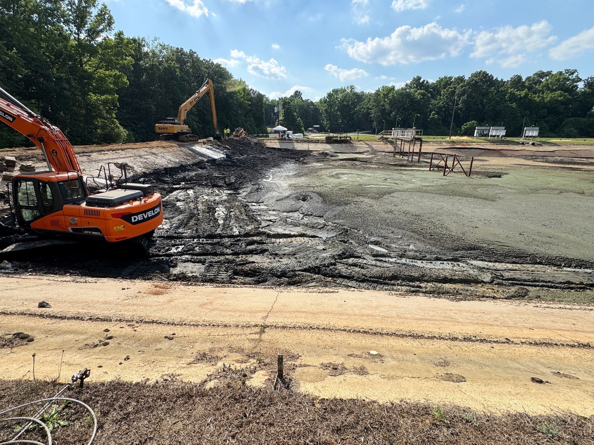 Tracked excavator and long-reach working solidified sludge with wastewater treatment plant visible during basin cleanout in eastern North Carolina