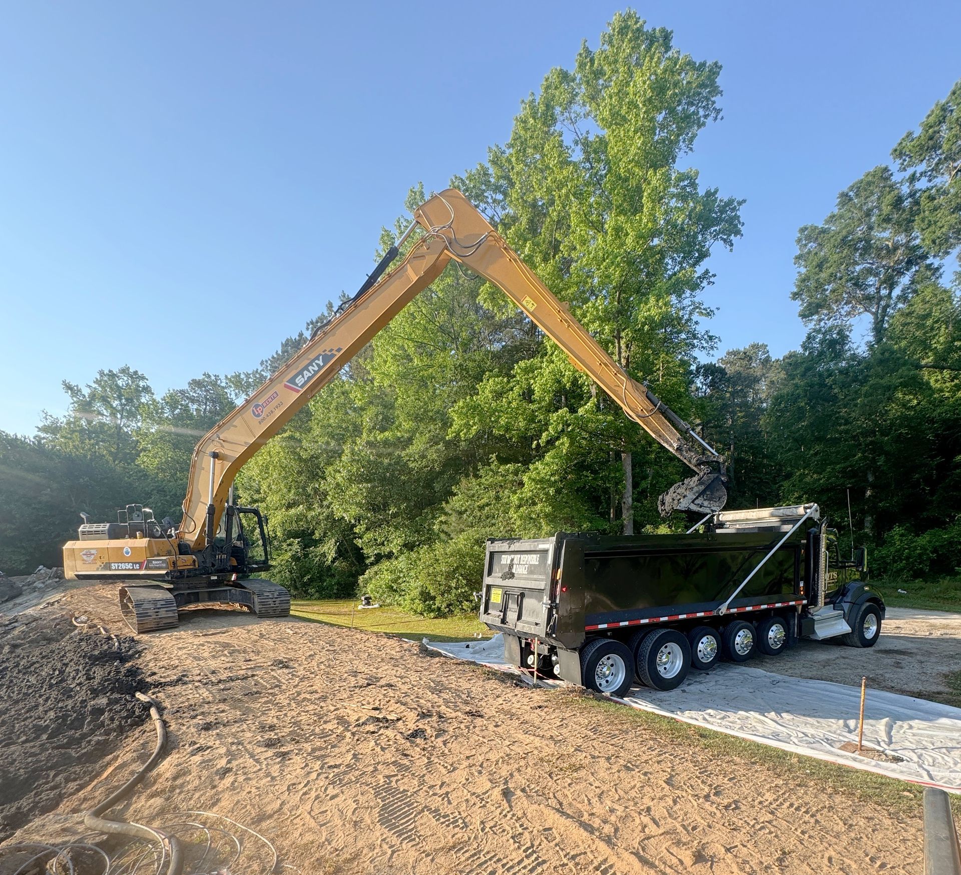 Long-reach excavator loading solidified sludge into a haul truck with broken concrete skirt debris staged alongside the basin in North Carolina