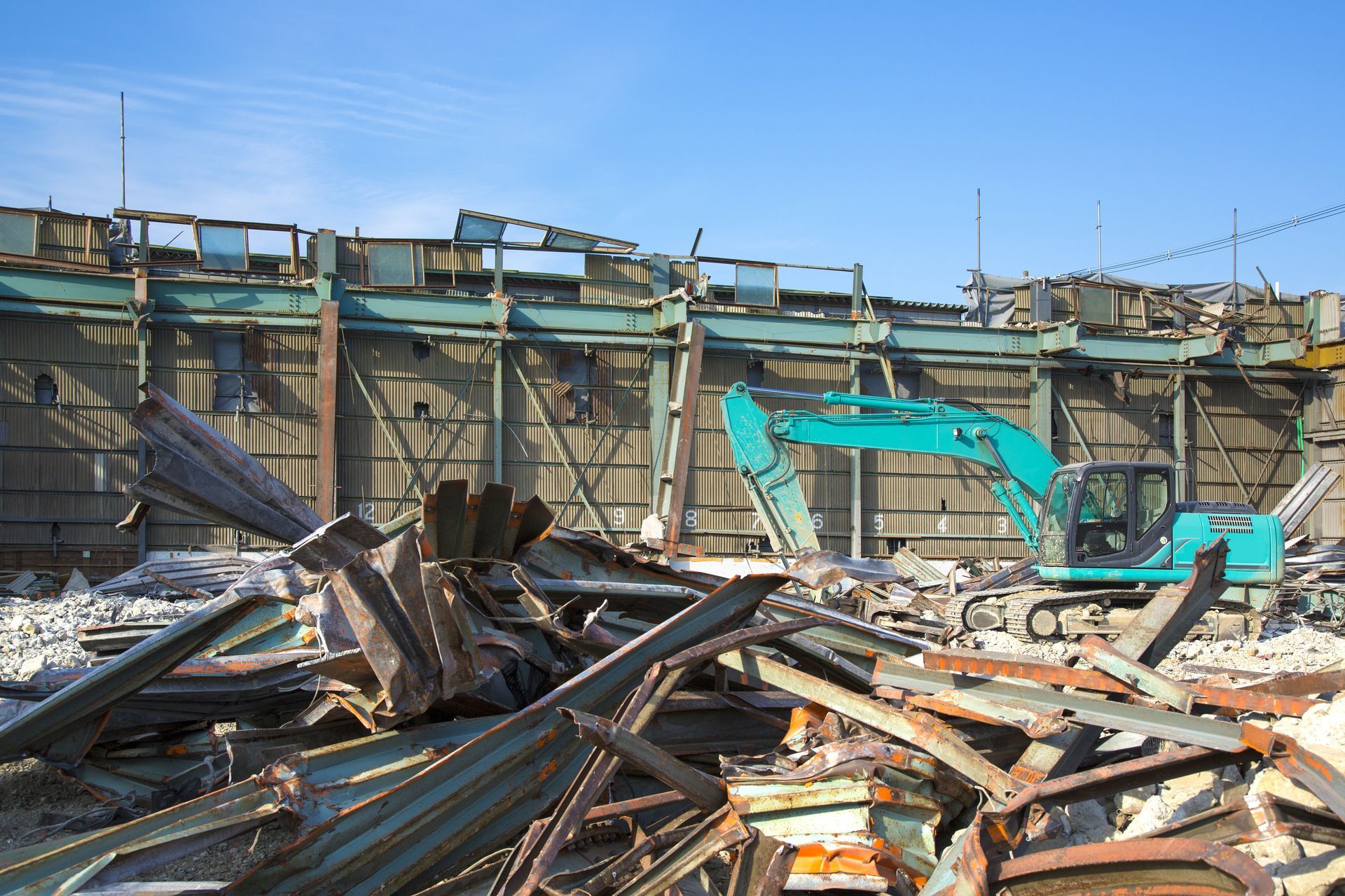 Collapsed building debris with twisted metal and a turquoise excavator in the background