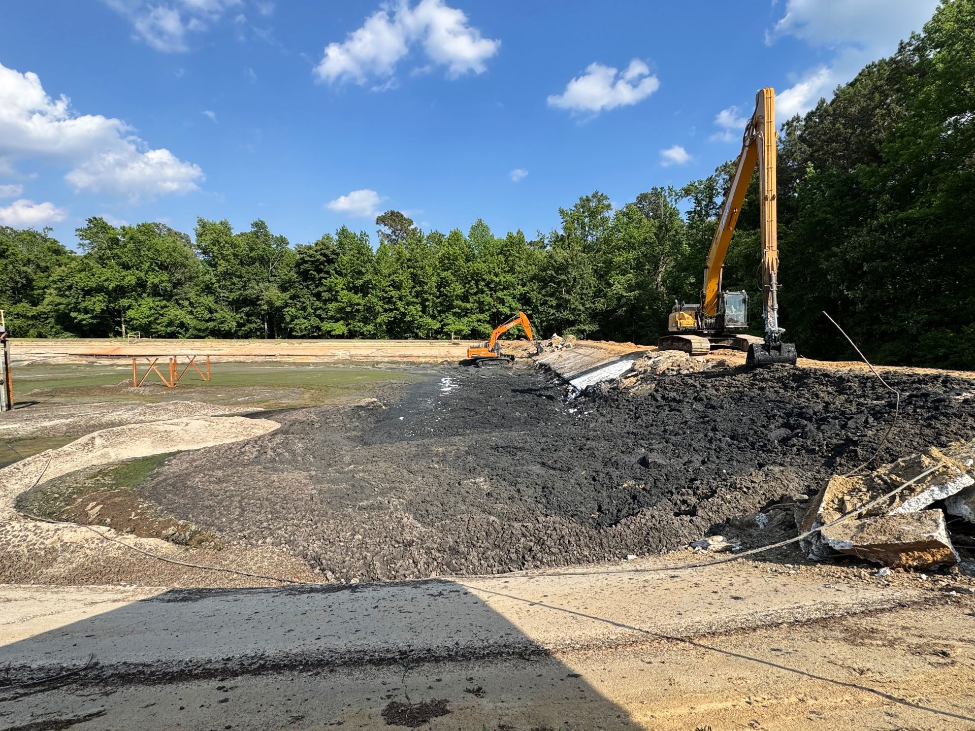 Long-reach and tracked excavator working solidified sludge with broken concrete skirt debris visible during basin cleanout in North Carolina