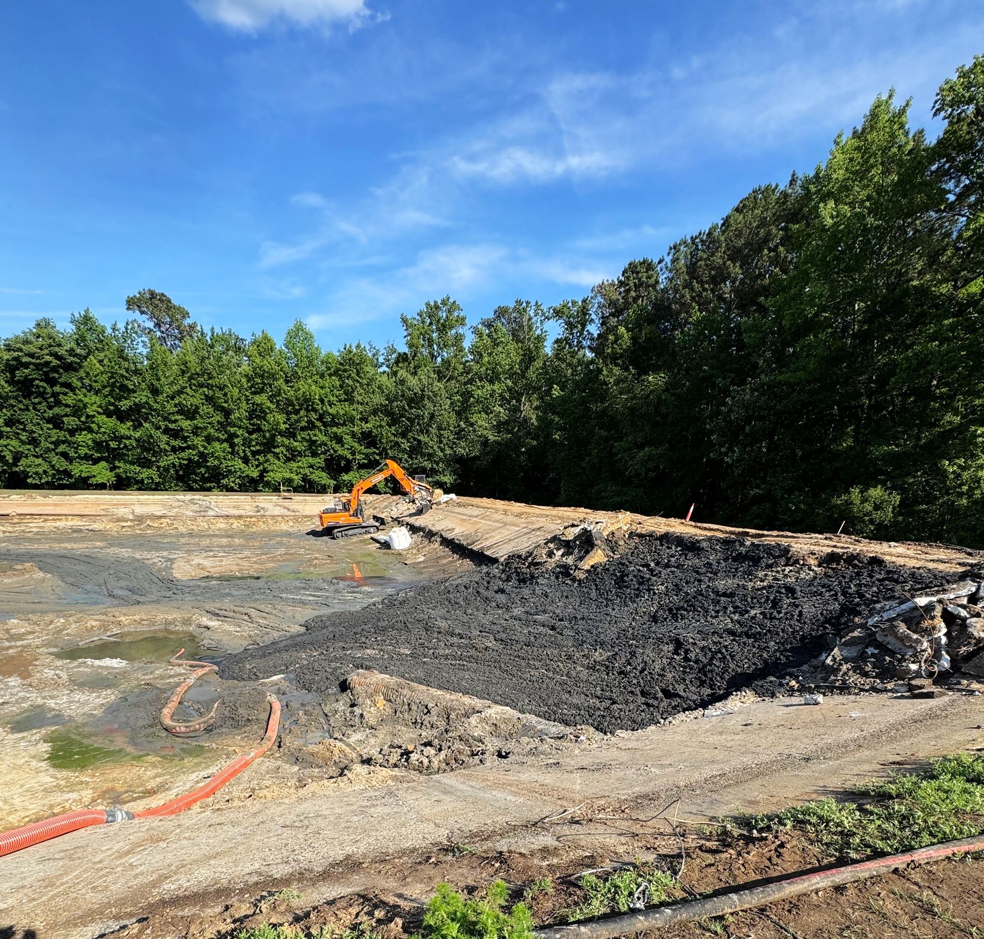 Tracked excavator working a solidified sludge wall with dewatering hoses running along the basin perimeter during North Carolina lagoon cleanout