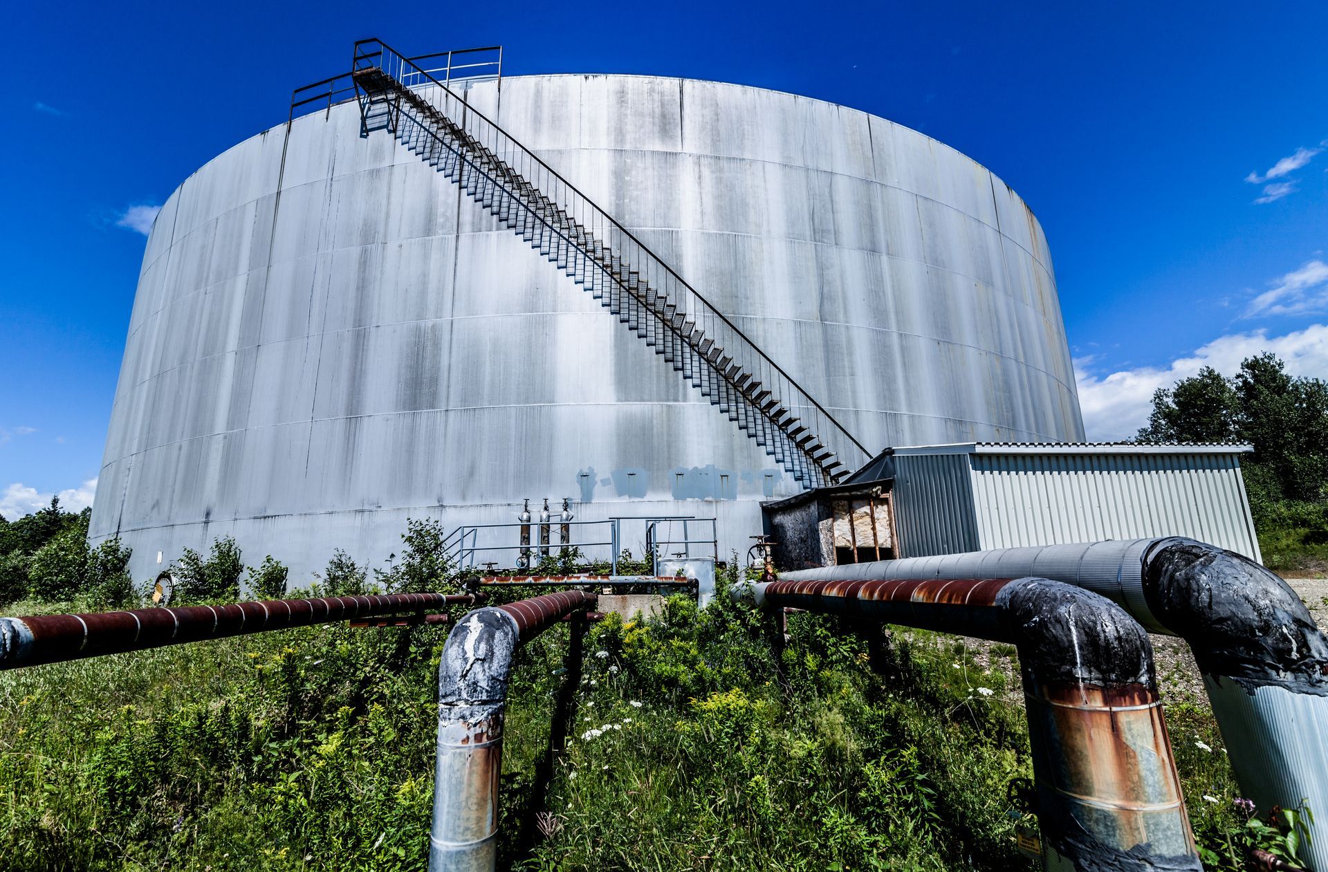 Large silver industrial storage tank with stairs and pipes under a bright blue sky