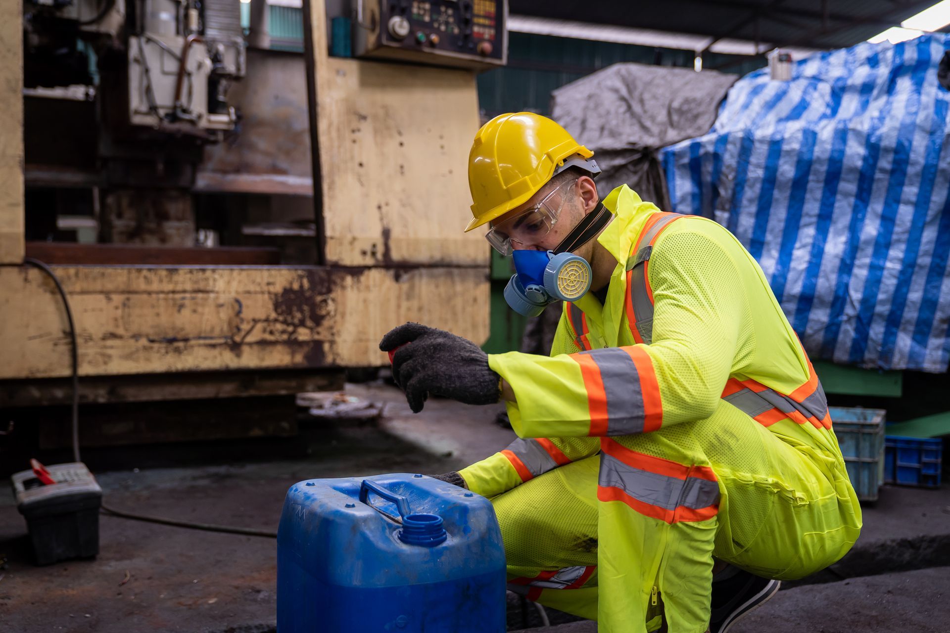 Worker in neon safety gear and helmet crouches by a blue container in an industrial setting.