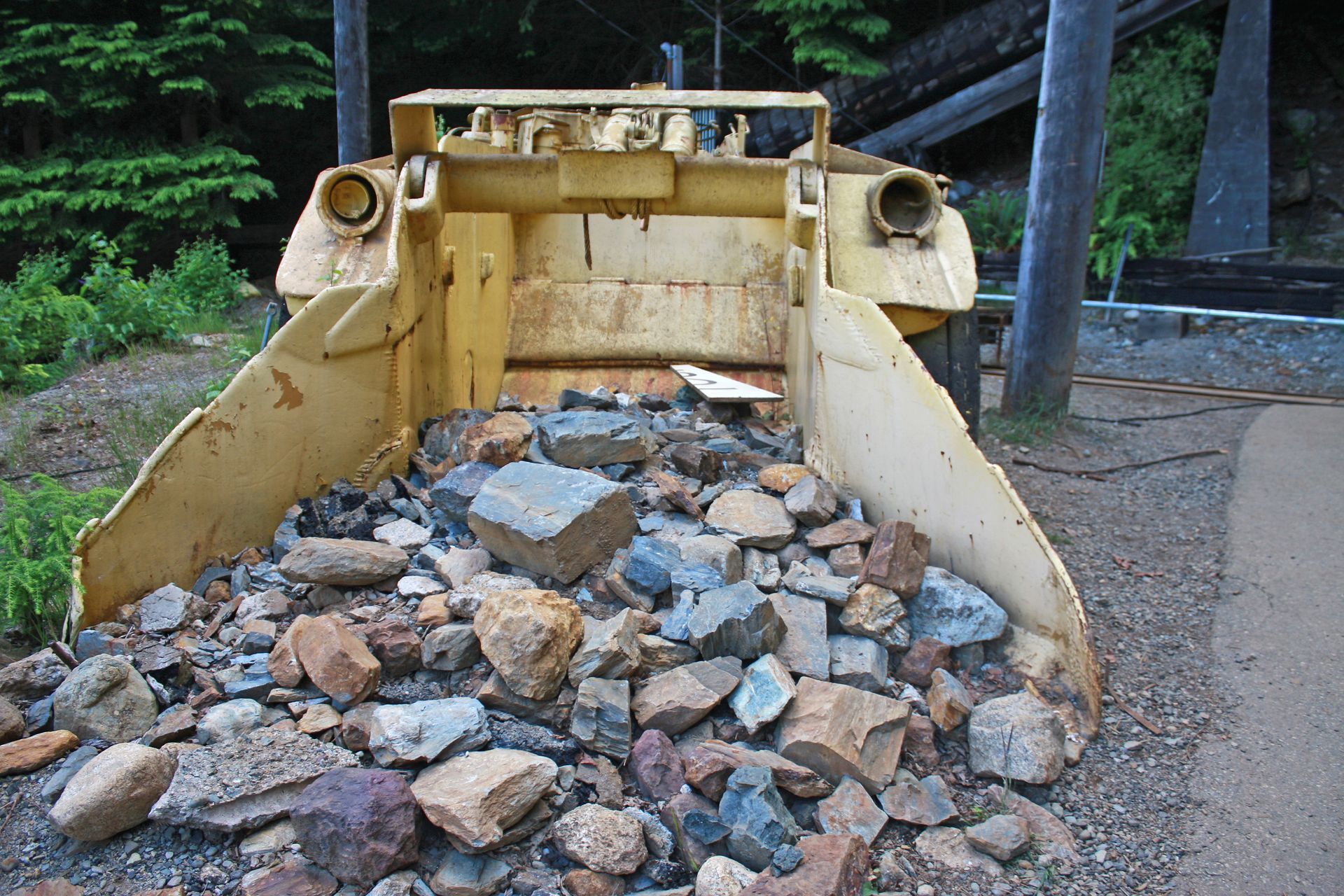 Yellow excavator bucket filled with rocks beside a dirt path and trees