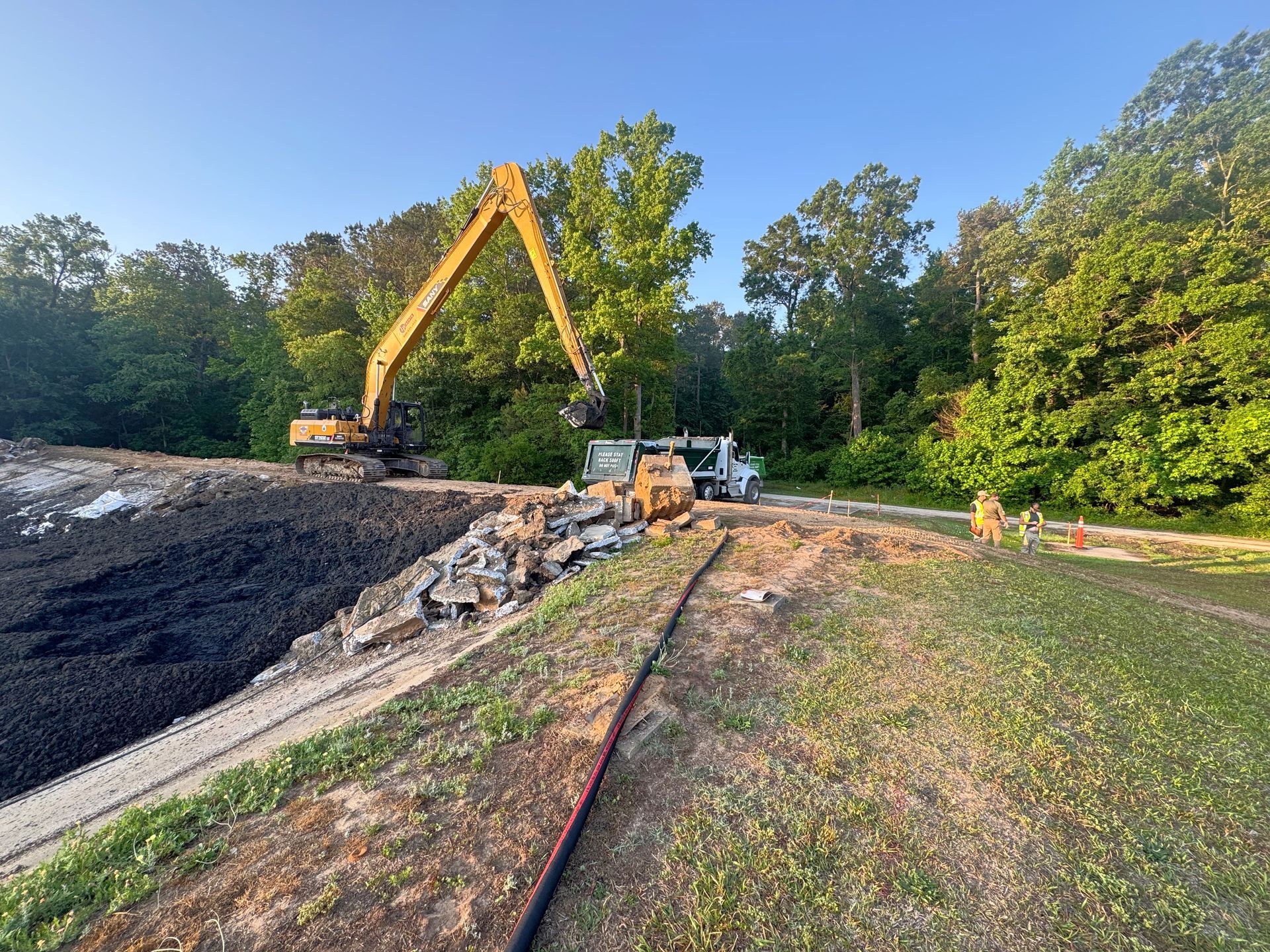 Long-reach excavator loading broken concrete skirt debris into a dump truck with crew members on site during North Carolina basin cleanout