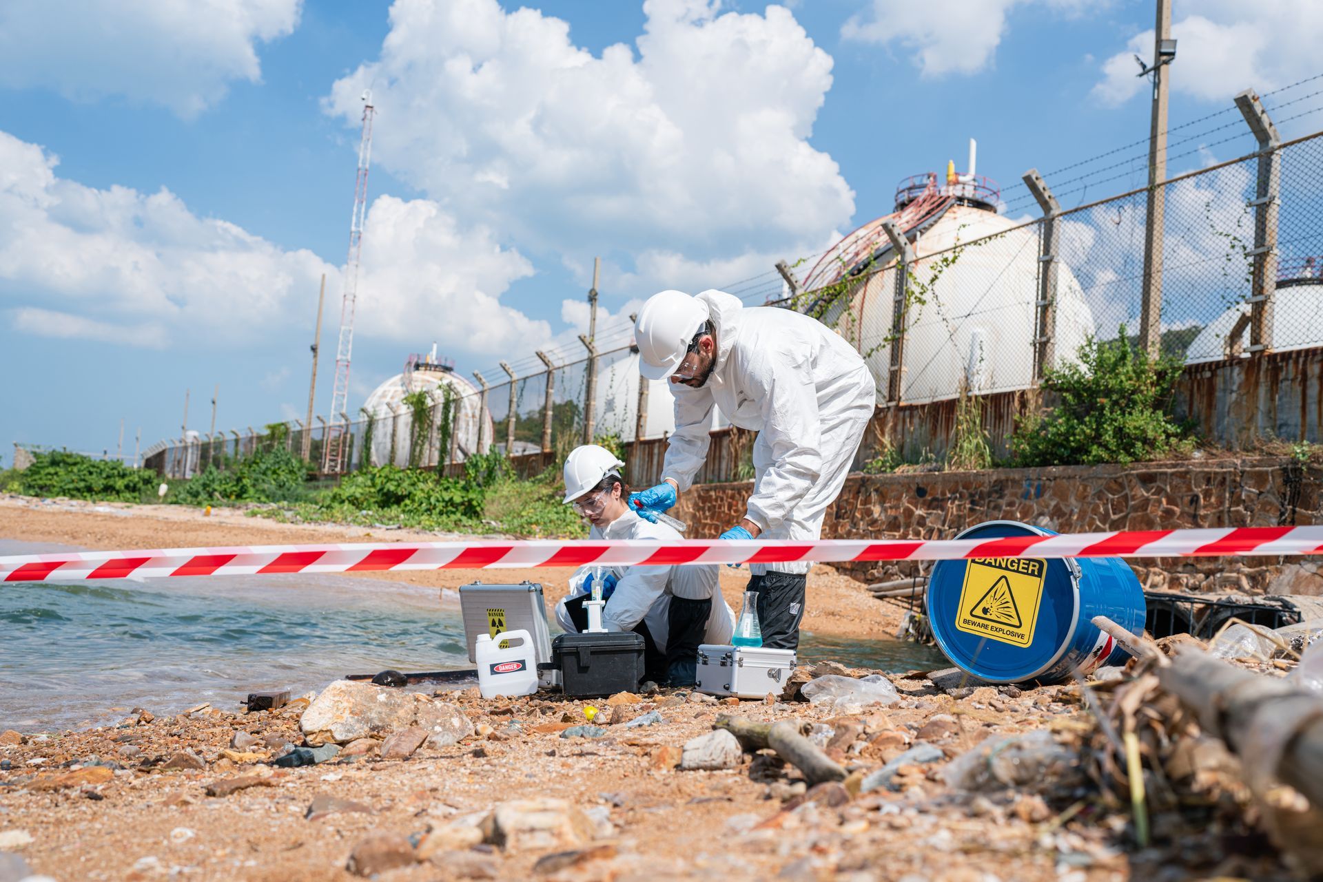 Workers inspecting debris behind red-and-white tape near industrial tanks on a roadside under a blue sky