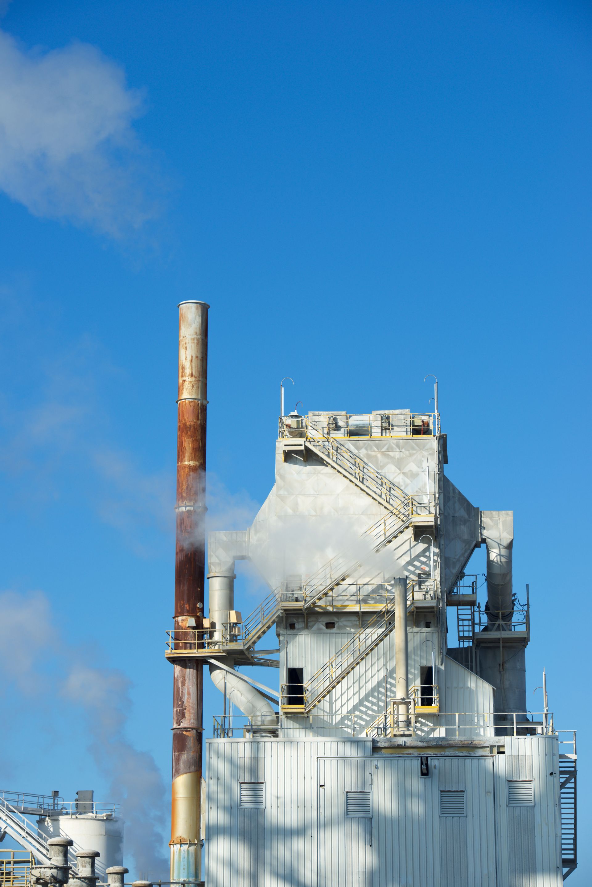 Industrial facility with white silos, a tall rusted smokestack, and blue sky with steam.
