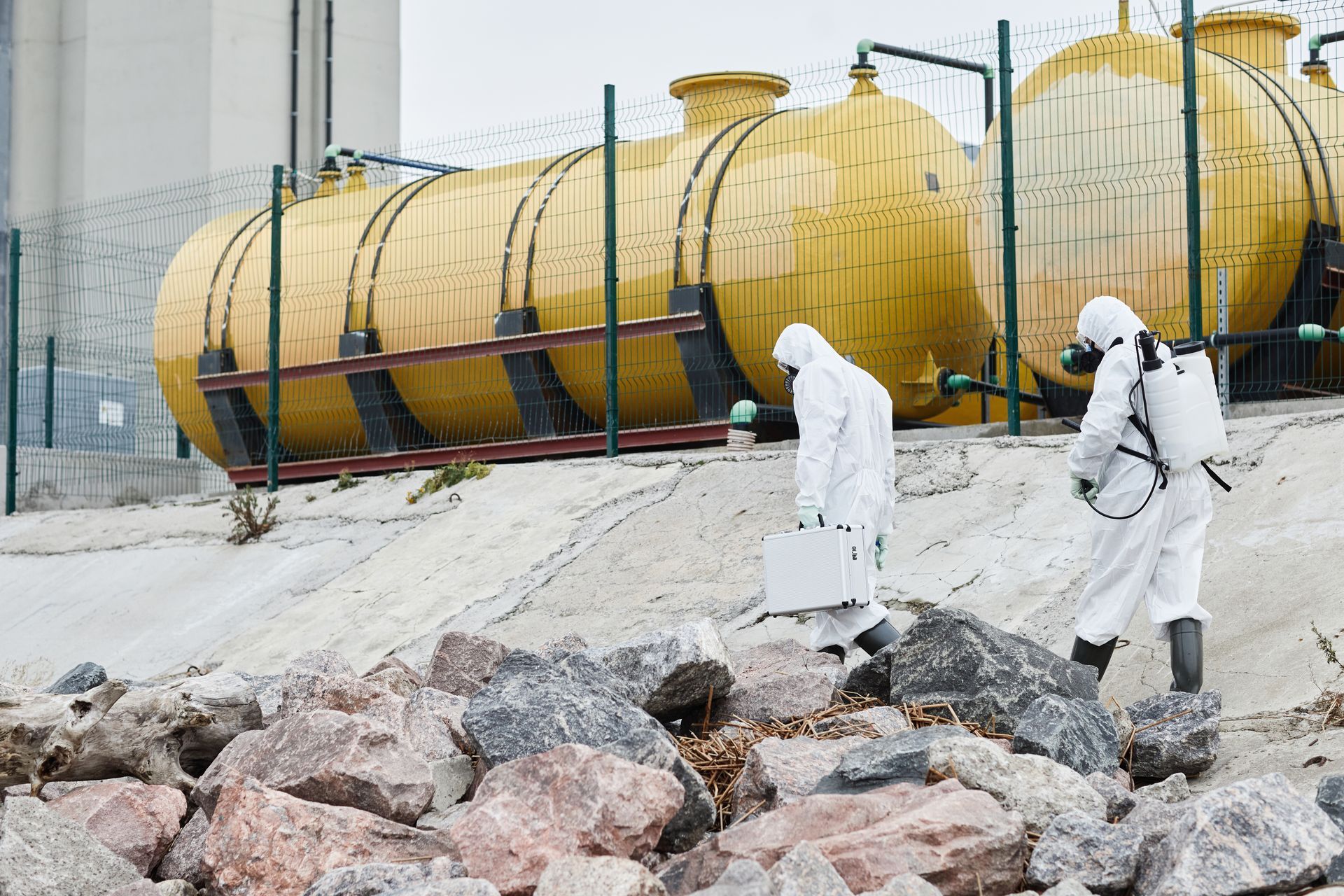 Workers in white protective suits inspect large yellow industrial tanks behind a chain-link fence
