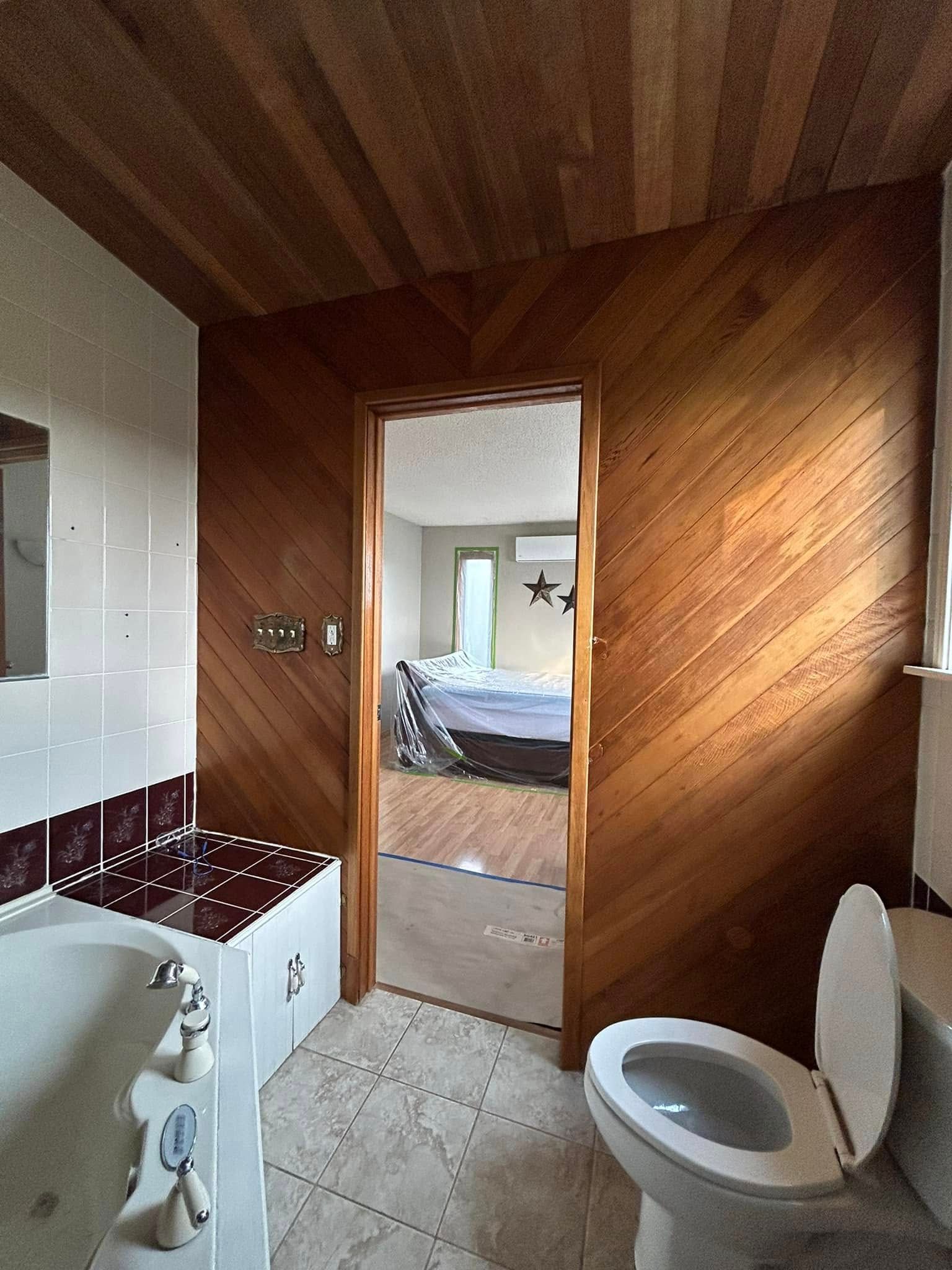 Before shot/outdated bathroom with wood paneled and white tile walls, toilet, bathtub, and doorway leading to a bedroom.