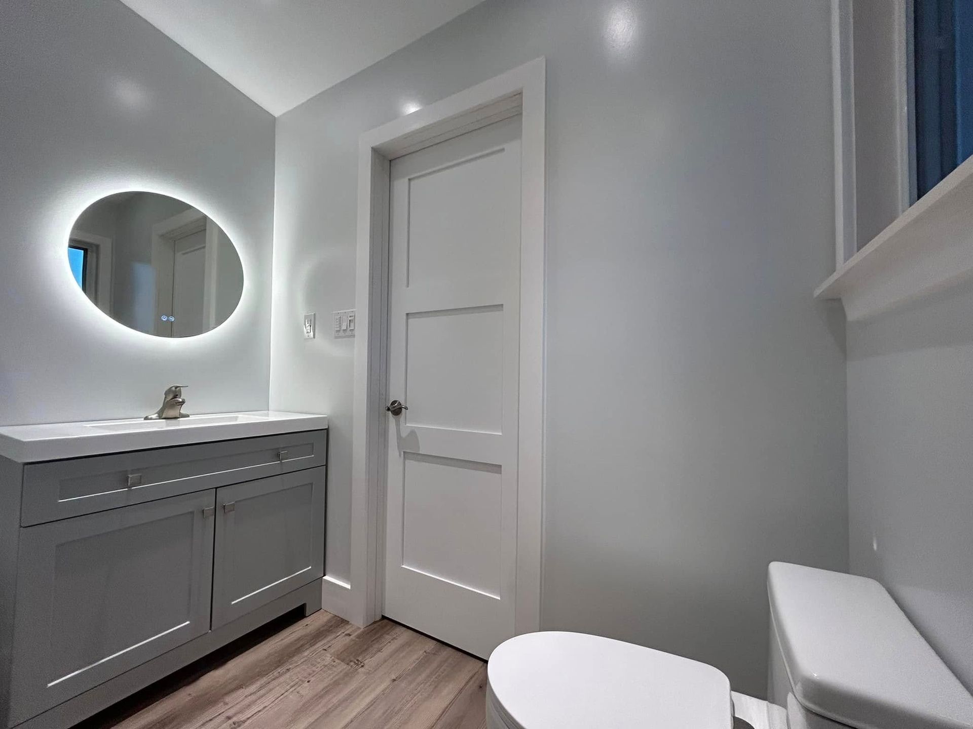 Gray bathroom with vanity, round lit mirror, door, and toilet.