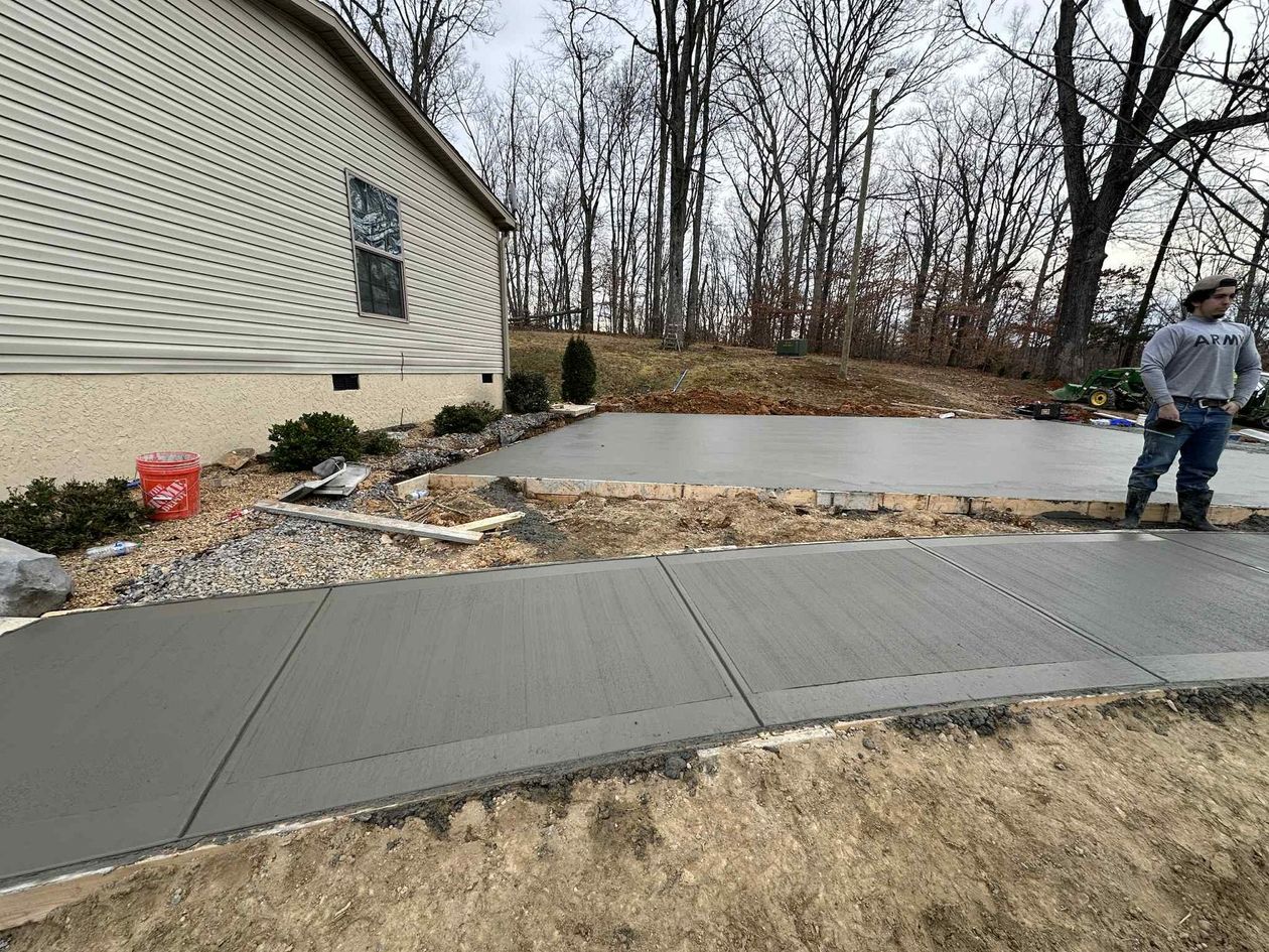 Concrete sidewalk and patio beside a house, with one person standing near the edge and winter trees in back.