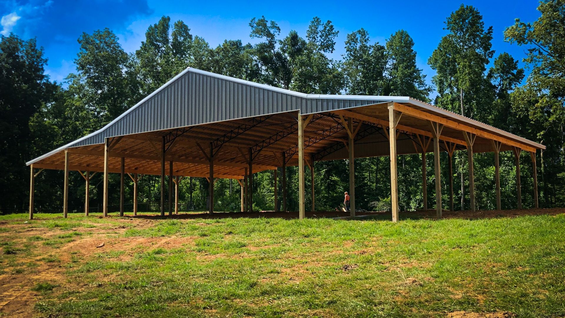 Open-air metal-roofed pavilion on a grassy field, supported by wooden posts. Trees in the background.