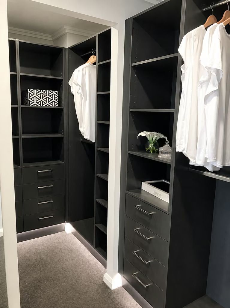 Dark walk-in closet with black shelving, hanging white clothes, drawers, and carpeted floor