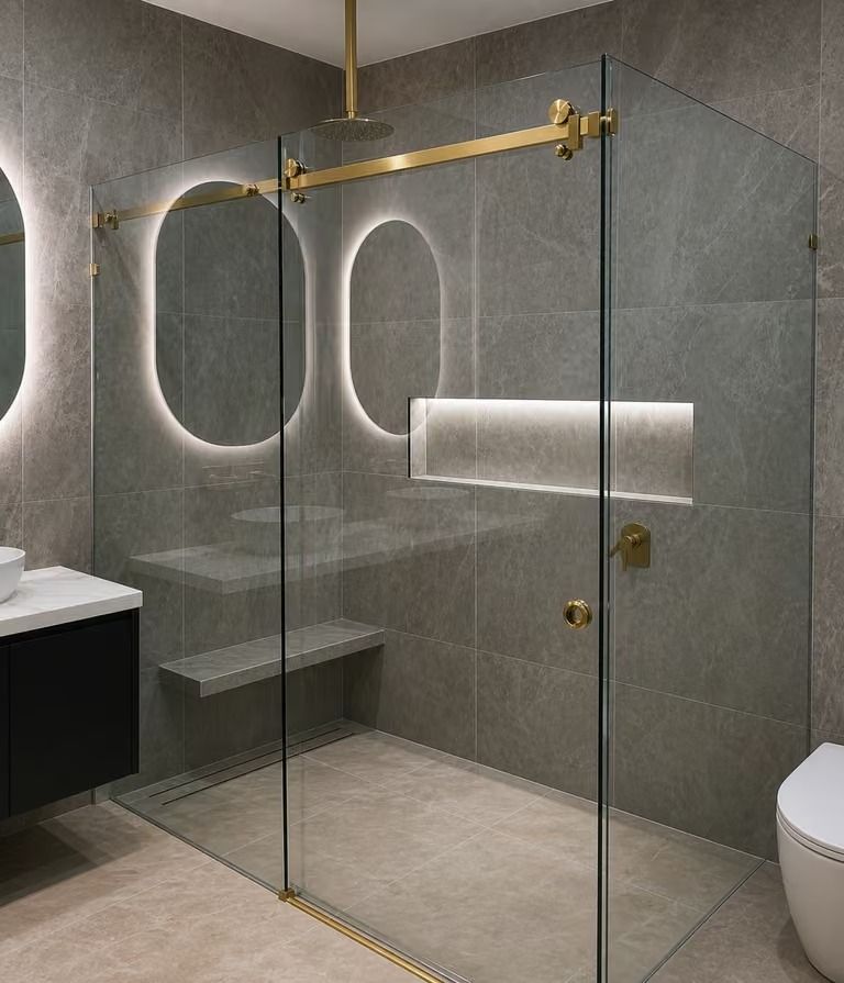 Modern bathroom with glass walk-in shower, brass fixtures, gray tile, and backlit oval mirrors