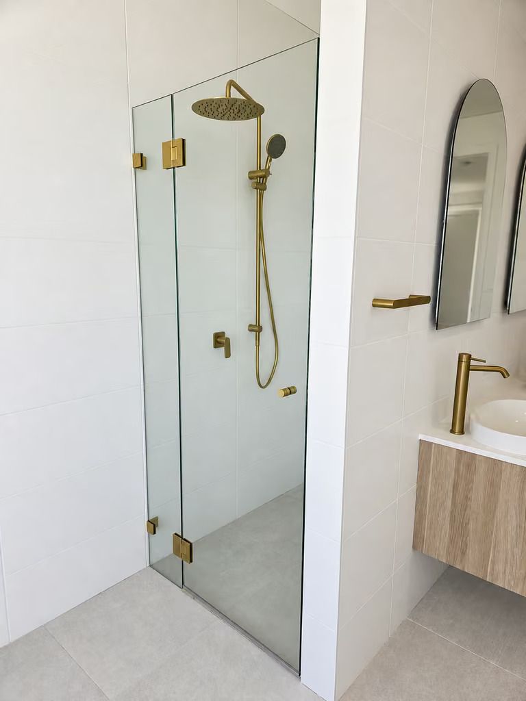 Corner shower with glass door, brass fixtures, and adjoining vanity with round mirror and gold faucet
