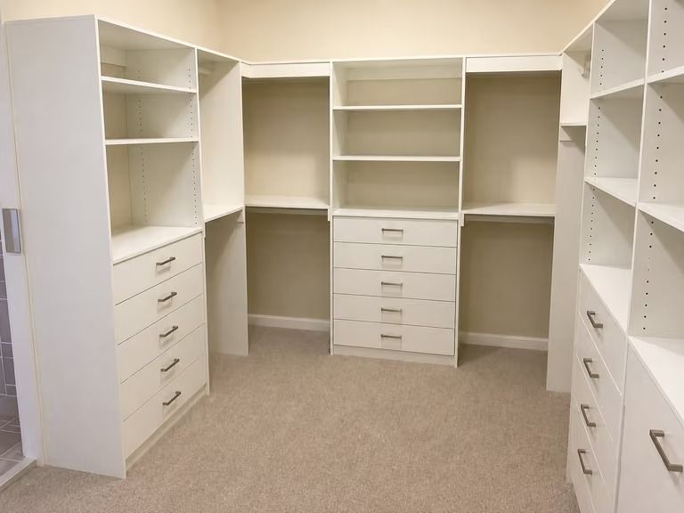 Empty walk-in closet with white shelves and drawers on beige carpet.