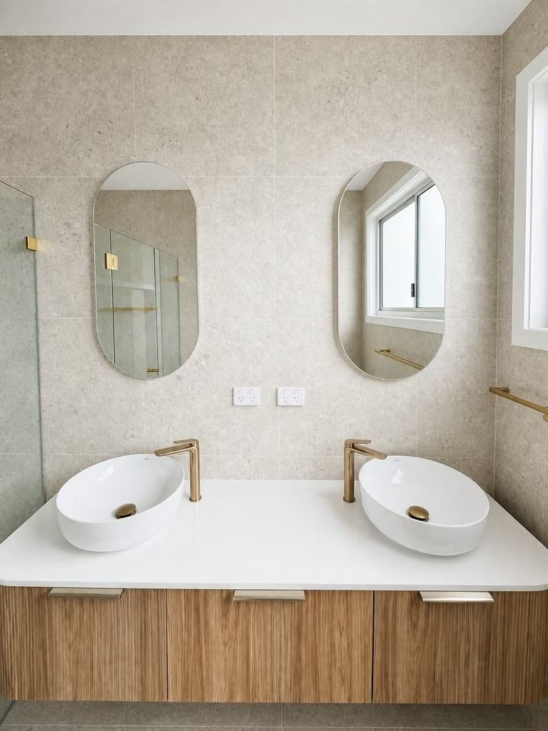 Modern bathroom vanity with two round sinks, brass faucets, oval mirrors, and wood cabinetry.