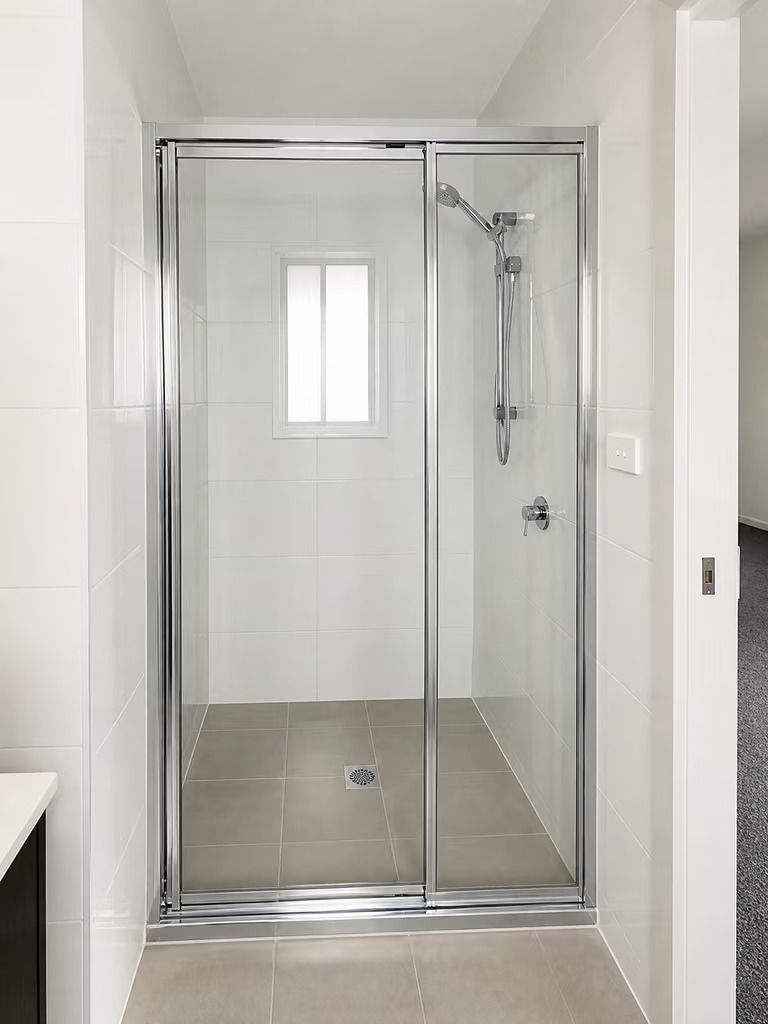 Glass shower enclosure in a white tiled bathroom with a handheld showerhead and gray floor tiles
