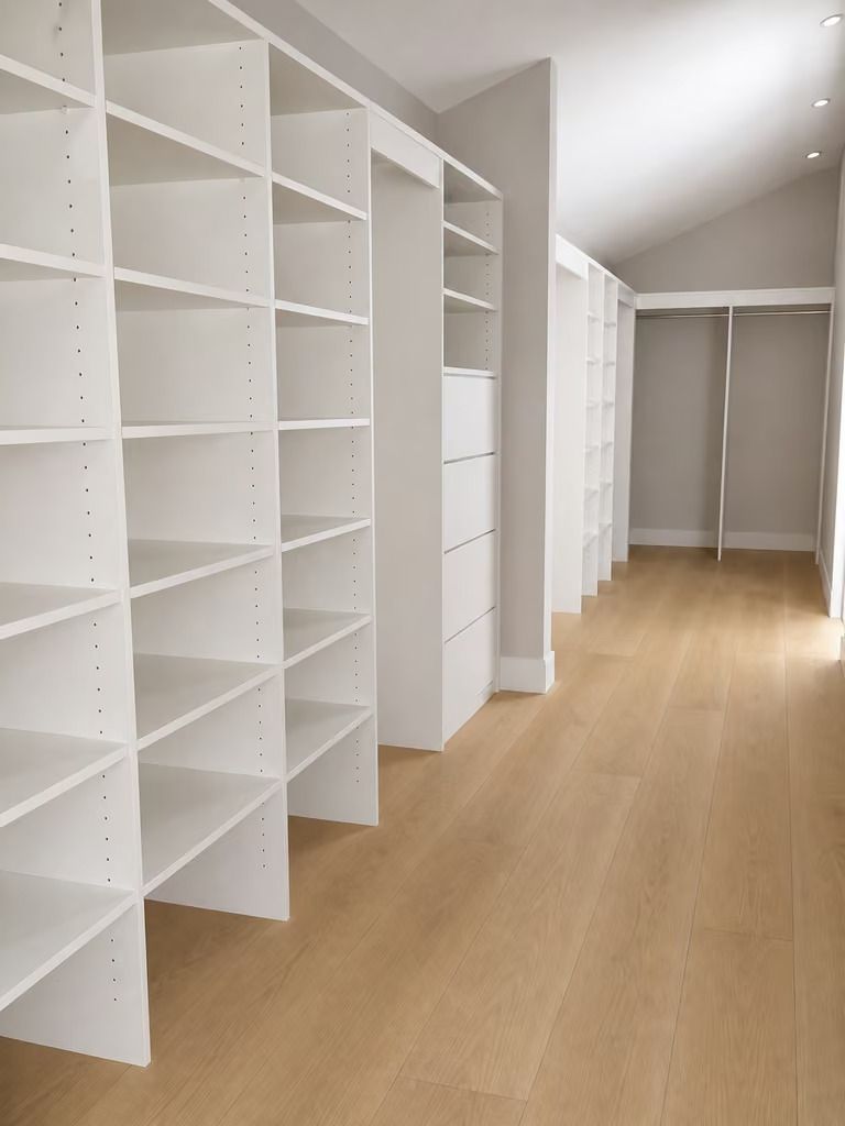 Bright walk-in closet with white built-in shelves and drawers along a light wood floor corridor