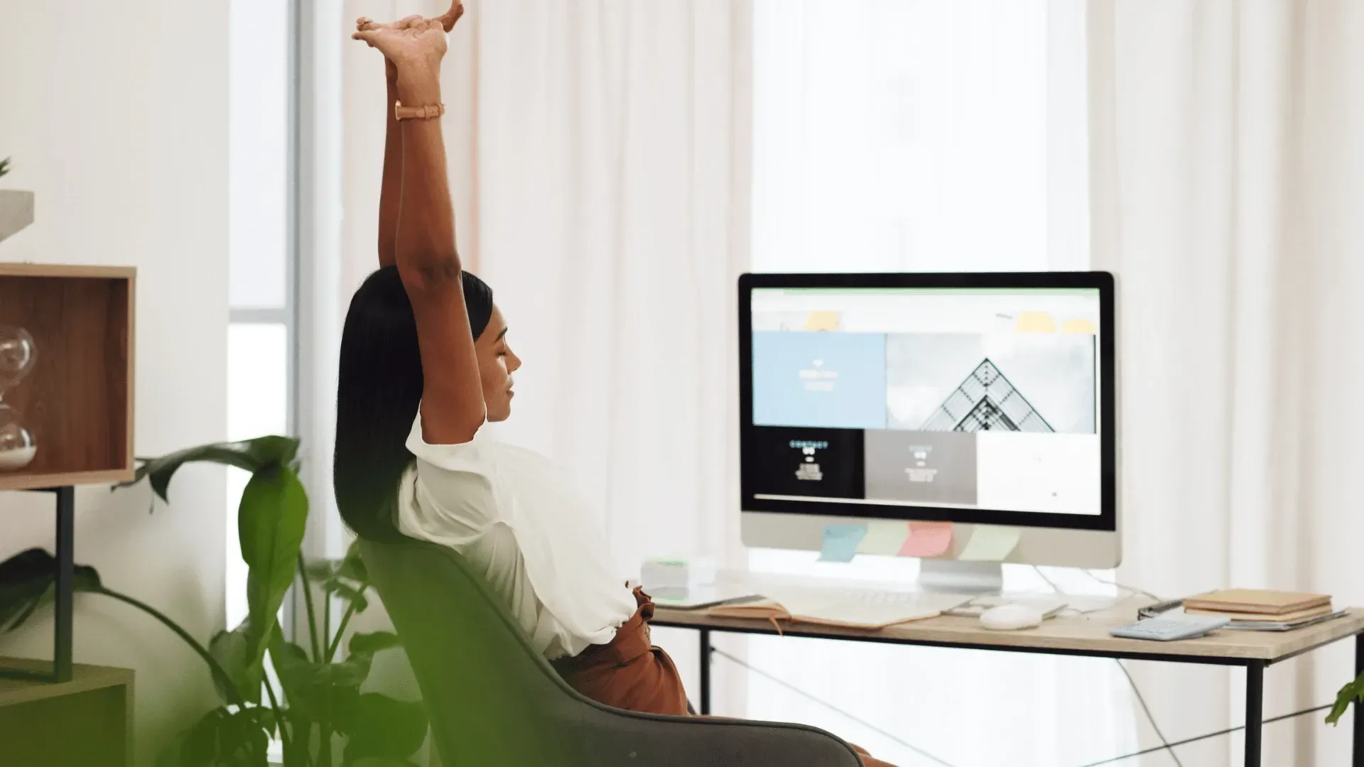 A woman is sitting at a desk stretching her arms in front of a computer.