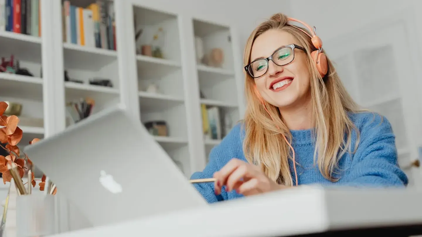Woman with blonde hair wearing glasses and headphones smiles while looking at a laptop. She is in a room with bookshelves.