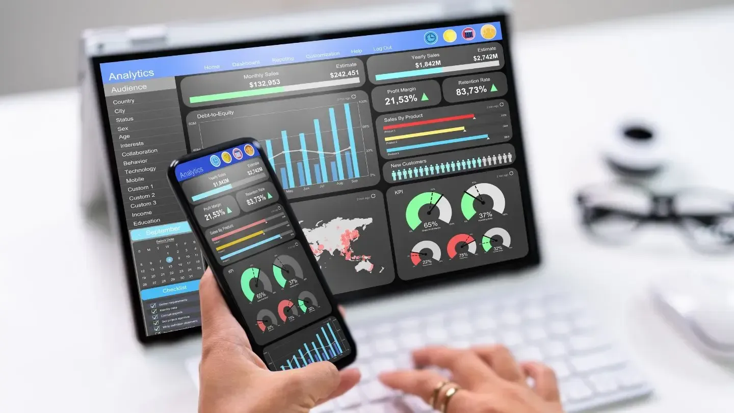 Person using a laptop and smartphone, both displaying data analytics dashboards. The screen shows colorful charts and graphs.