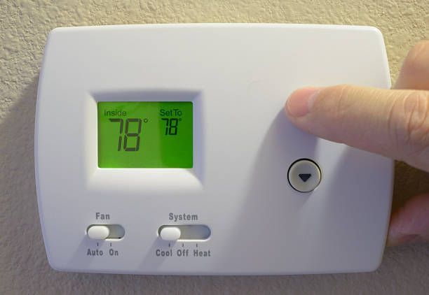 Hand pressing a button on a white, modern thermostat mounted on a light wood-paneled wall.