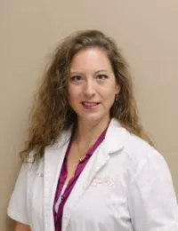 A woman in a white lab coat and purple tie is standing in front of a wall.
