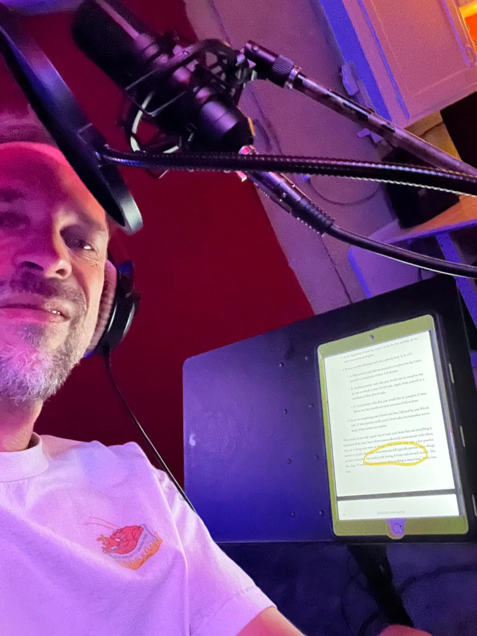 Man wearing headphones beside a microphone, reading from a glowing phone on a stand in a red-lit studio