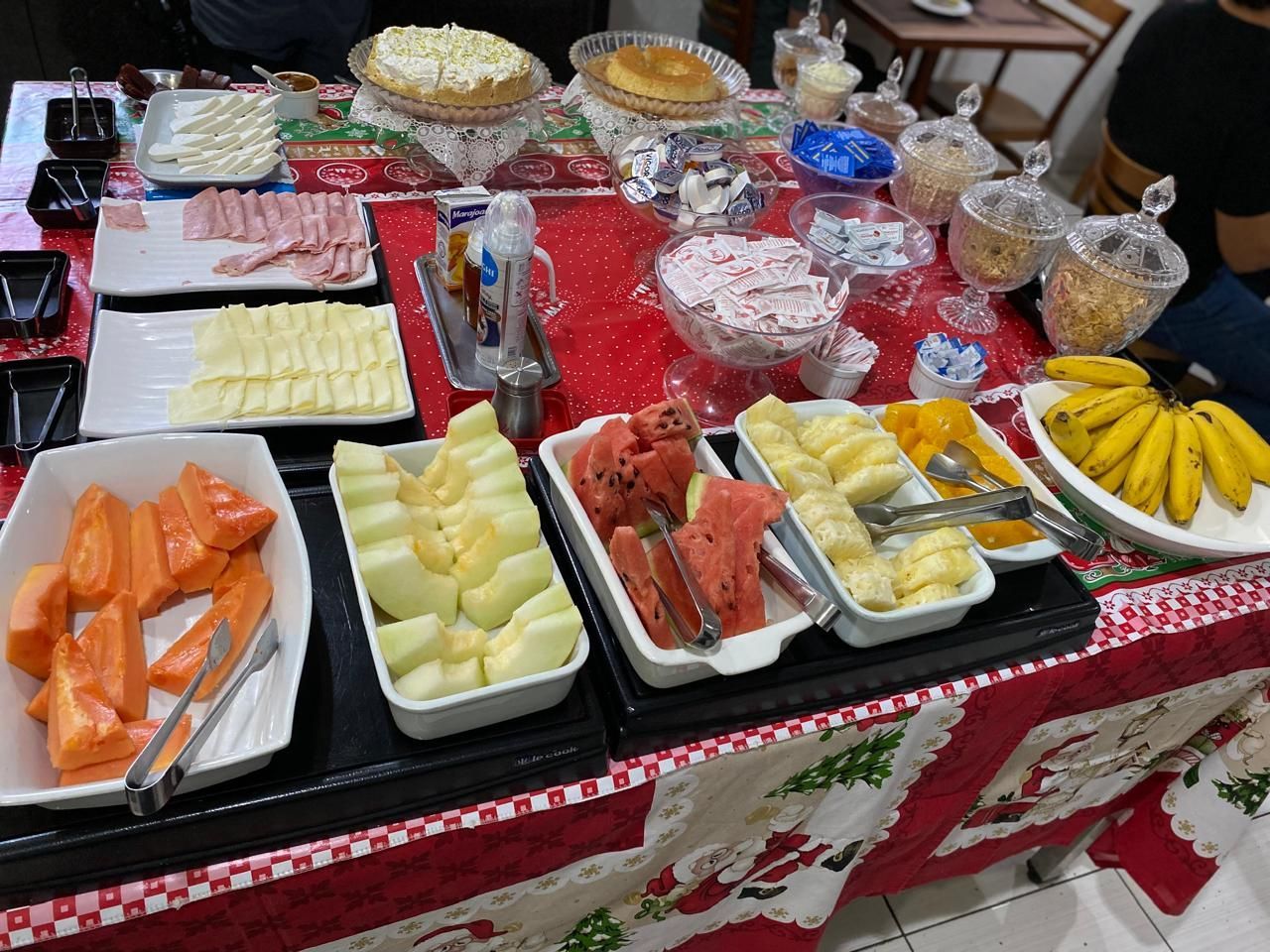 Mesa de bufê com bandejas de frutas variadas, sobremesas e bebidas sobre uma toalha de mesa vermelha.