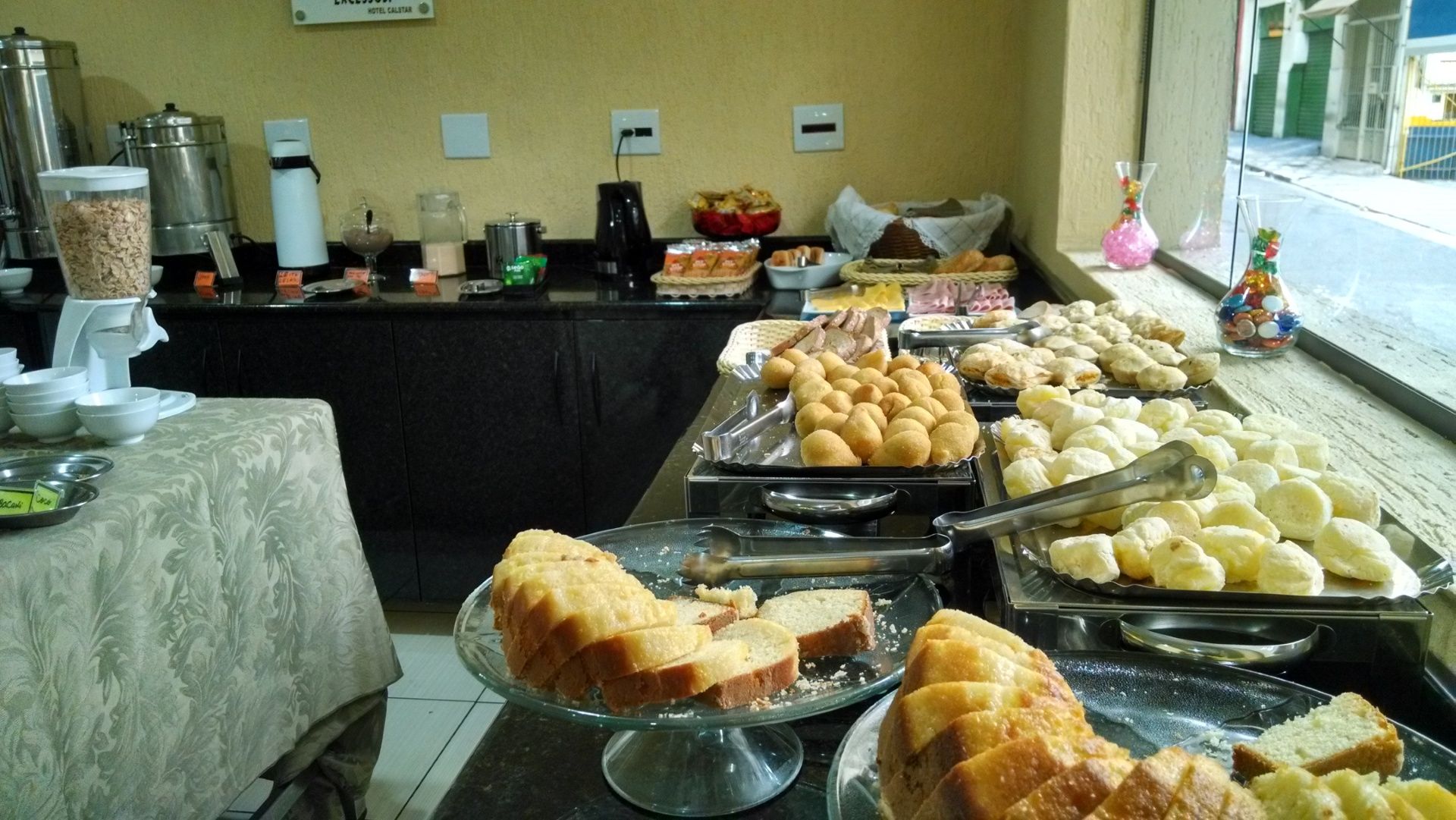 Buffet de café da manhã com doces, pãezinhos, frutas e café em um balcão junto à janela.