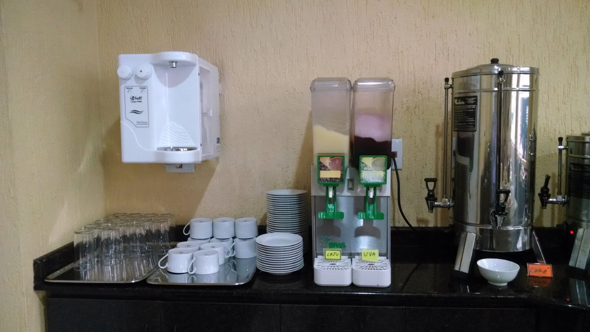Estação de café e chá com xícaras, dispensadores e uma grande chaleira sobre uma bancada contra uma parede bege.
