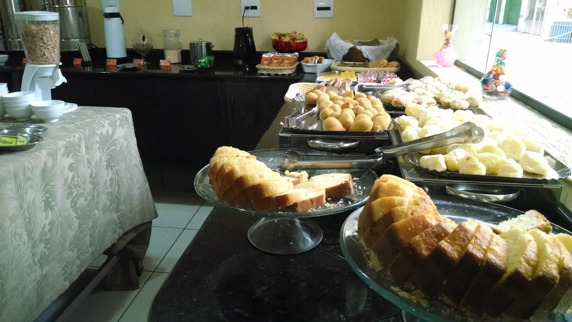 Mesa de buffet com bandejas de doces variados e fatias de bolo em uma sala de jantar iluminada.